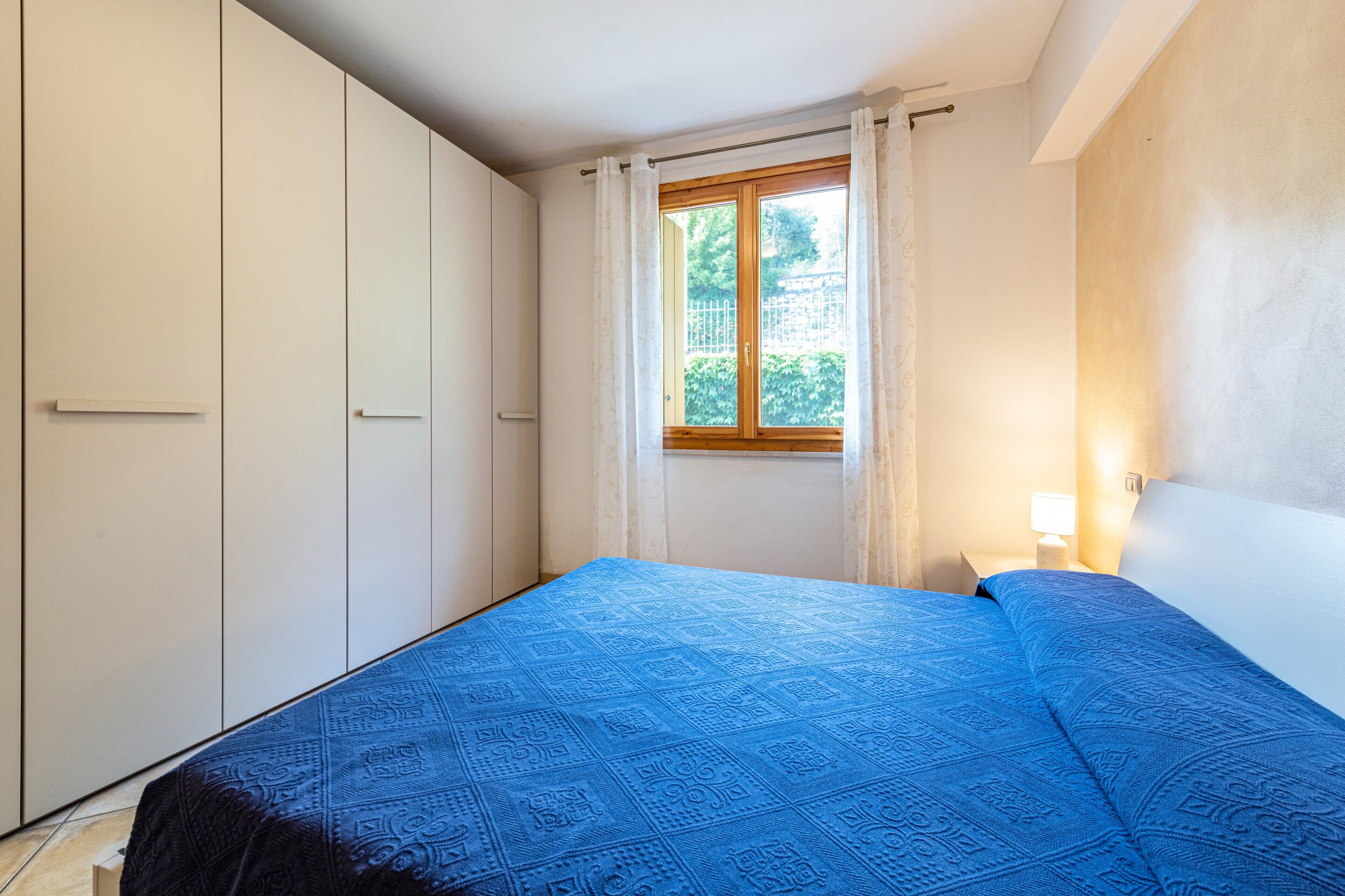 A bedroom with a bed, a window with curtains, a bedside lamp, and a wardrobe.