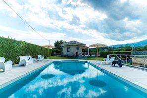 Una piscina con sillas de descanso y sombrillas, un edificio con techo abovedado y montañas al fondo.