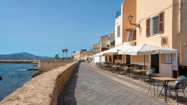 A seaside promenade with outdoor seating, a stone wall, and buildings with balconies.