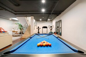 Une table de billard avec les billes disposées en pyramide, un escalier et une plante dans une pièce aux murs blancs et au plafond où les tuyaux sont apparents.