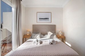 Une chambre d’hôtel avec un lit, deux tables de chevet, un tableau encadré au mur et une vue par la fenêtre.