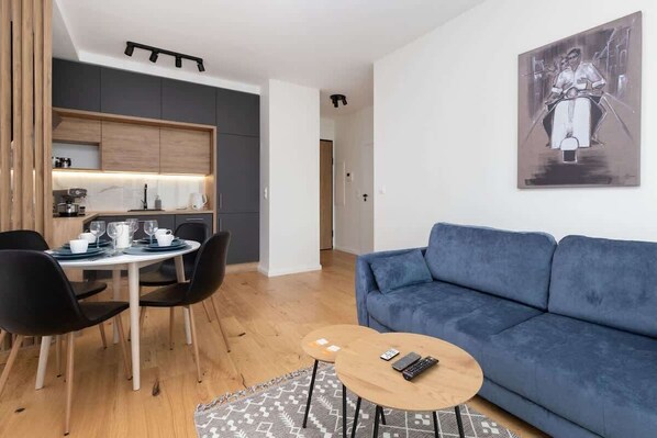 Un salon moderne avec un canapé bleu, un coin repas comprenant une table ronde et des chaises, et une cuisine avec des meubles sombres et un plan de travail clair.