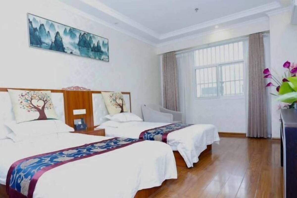 A hotel room with two beds, a painting on the wall, a window with curtains, and a vase with flowers on a table.