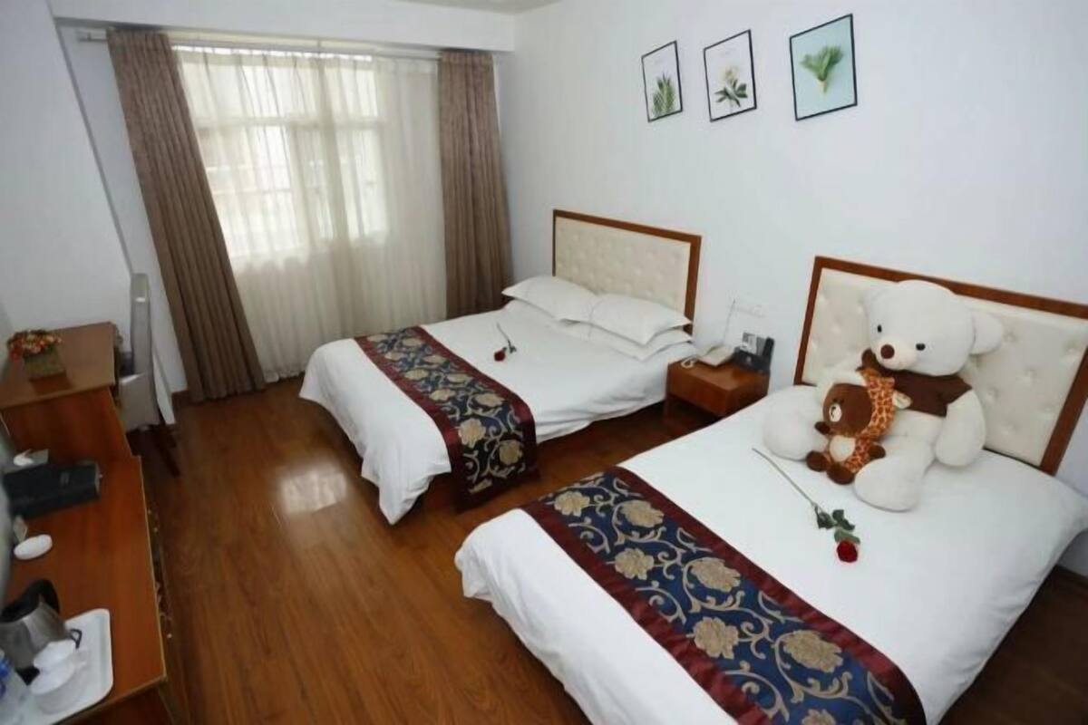 A hotel room with two beds, a wooden floor, a desk with a chair, and a window with curtains.