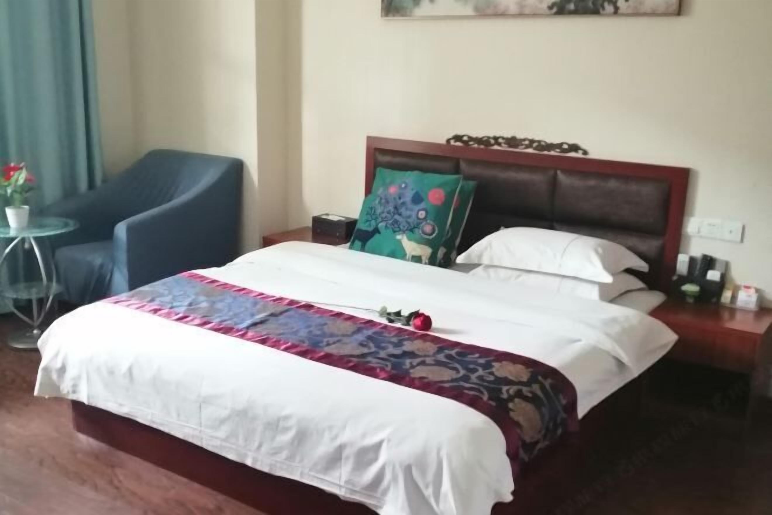 A neatly made bed with a floral bedspread, a blue armchair, and a small round table with a potted plant.