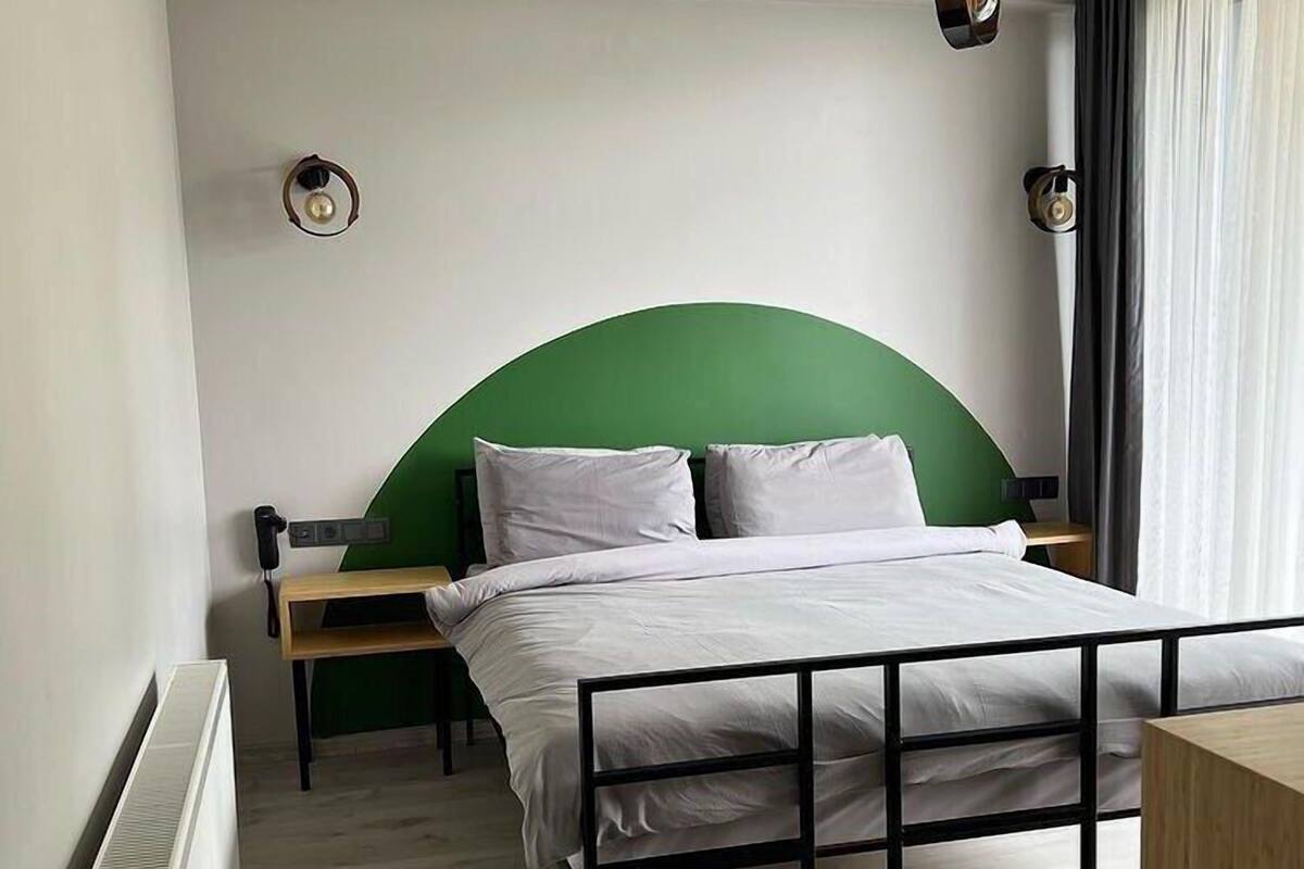 A bedroom with a green headboard, white bedding, a wooden nightstand, a bedside table, a radiator, and a window with curtains.