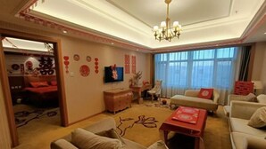 A well-lit living room with a sofa set, a coffee table, and a television. There is a window with curtains, a chandelier, and a door leading to another room. - Vienna International Hotel (Jilin Guotou Zhongdong) (Jilin City)