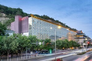 Un hôtel moderne à la façade en verre, situé près d’un bâtiment traditionnel et d’une rue animée.