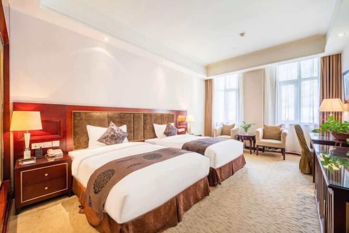 Quarto de hotel com duas camas, uma mesa de cabeceira, uma cadeira, uma escrivaninha e uma janela grande.