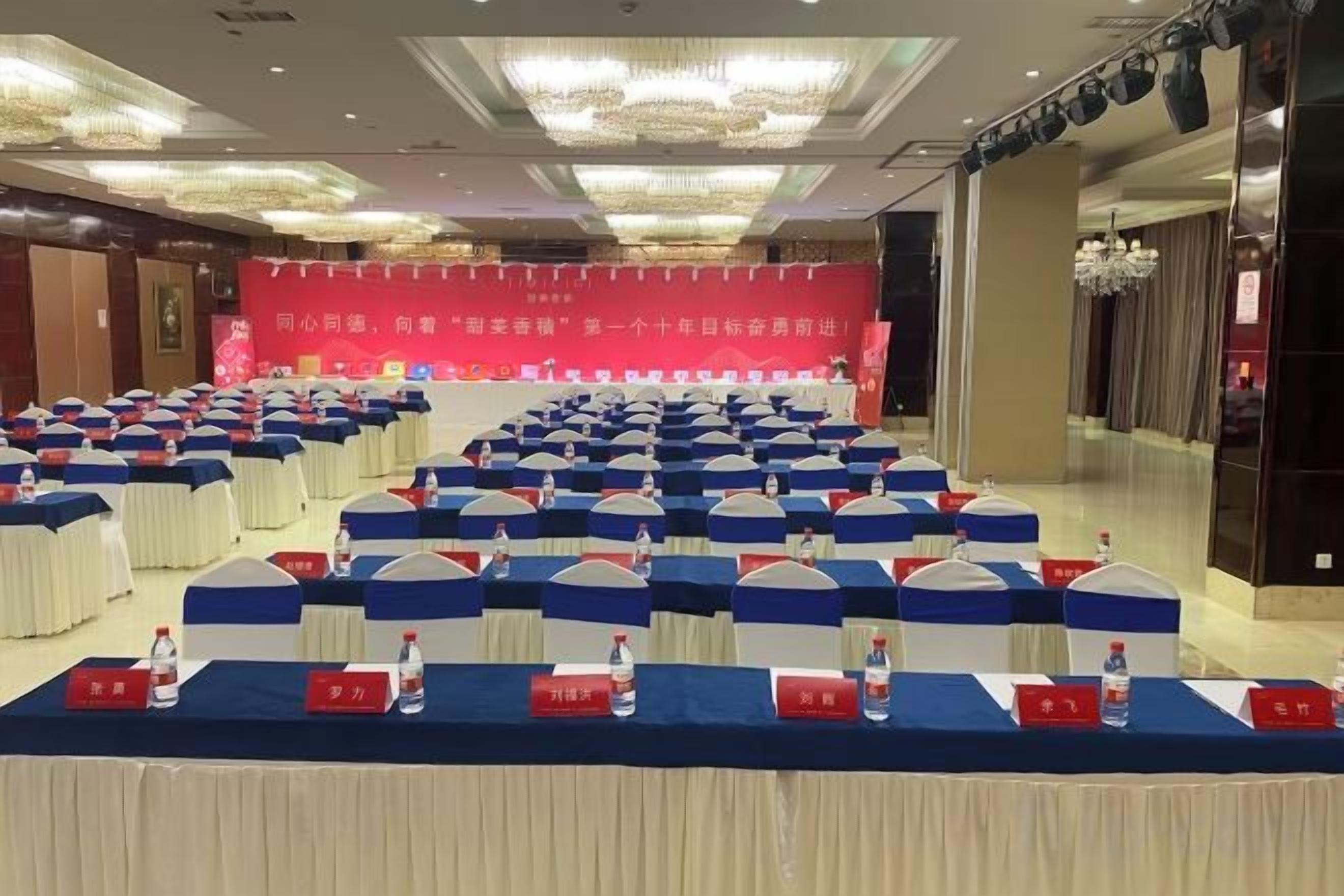 Une salle de conférence avec des rangées de tables, de chaises et des plaques nominatives, une scène ornée d’une banderole et des lustres.