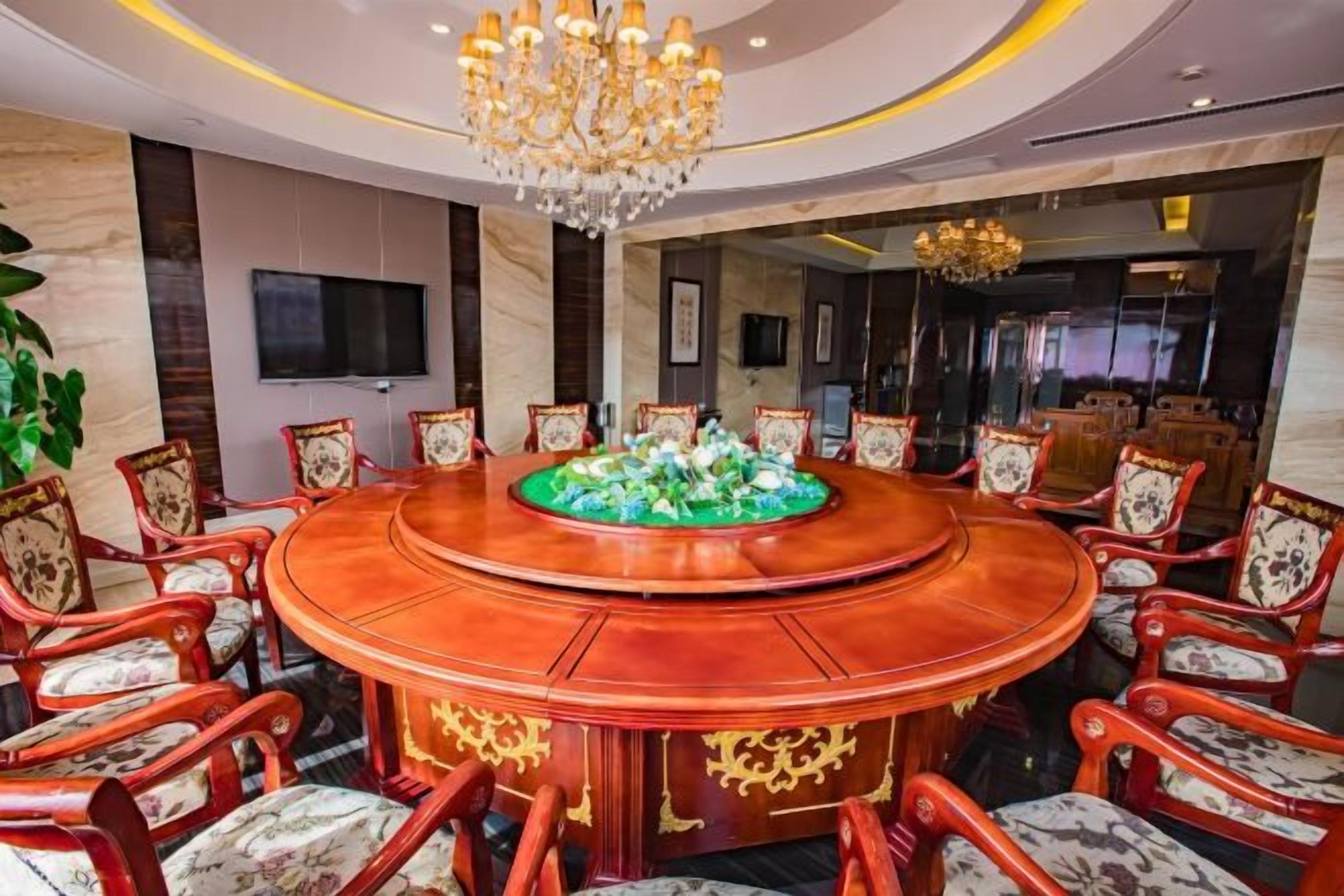 A conference room with a round table surrounded by chairs, a chandelier, and a television on the wall.