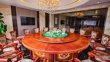 A conference room with a round table surrounded by chairs, a chandelier, and a television on the wall.
