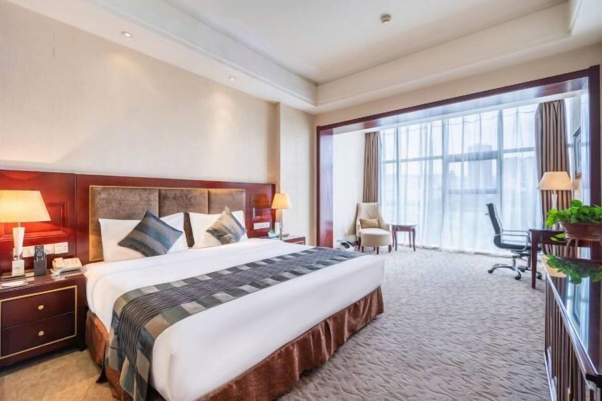 Quarto de hotel com uma cama grande, uma mesa de cabeceira, uma cadeira, uma escrivaninha e uma janela ampla.