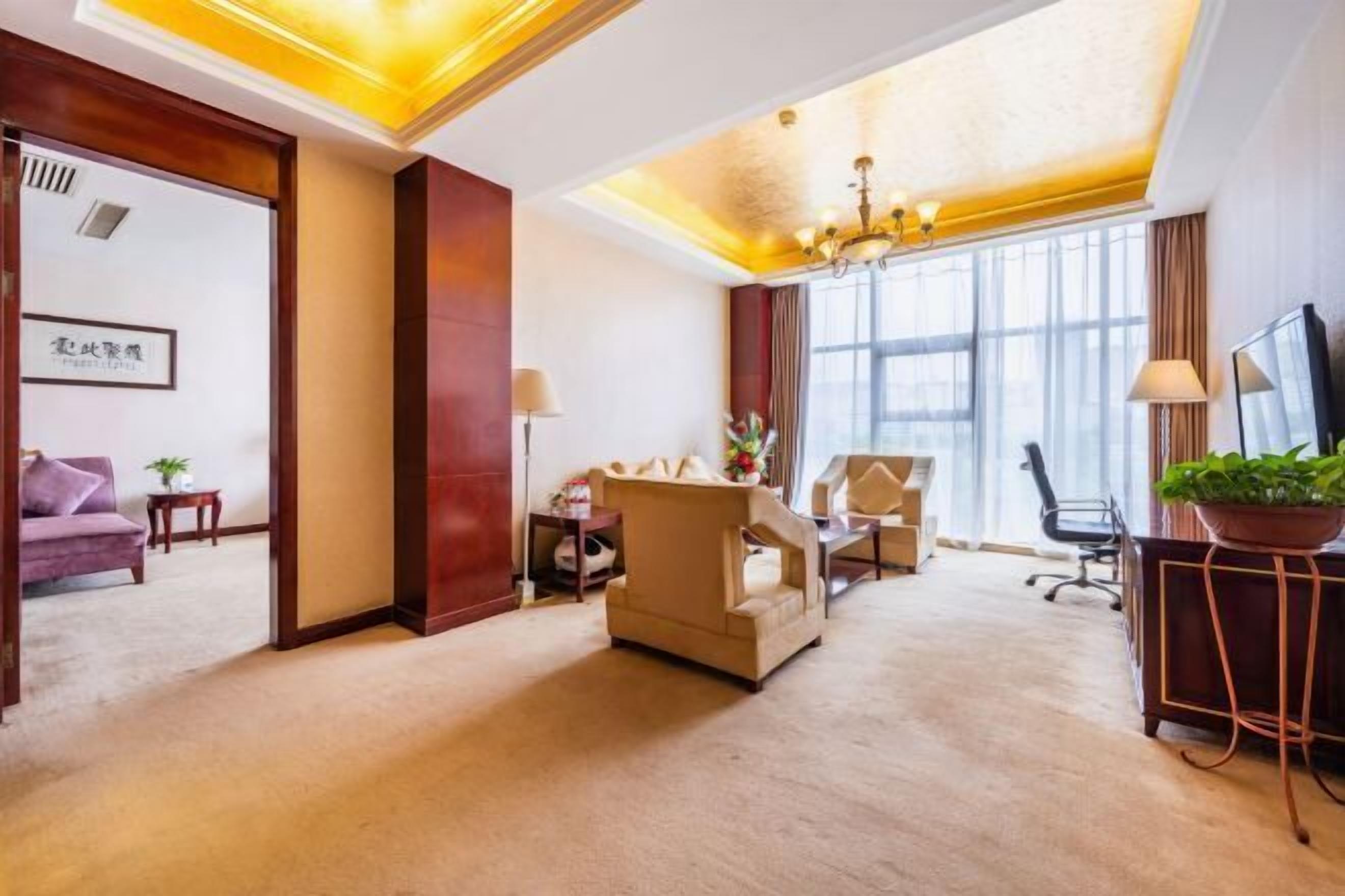 Une chambre d’hôtel avec une grande fenêtre, un lustre, un canapé, un bureau et une télévision.