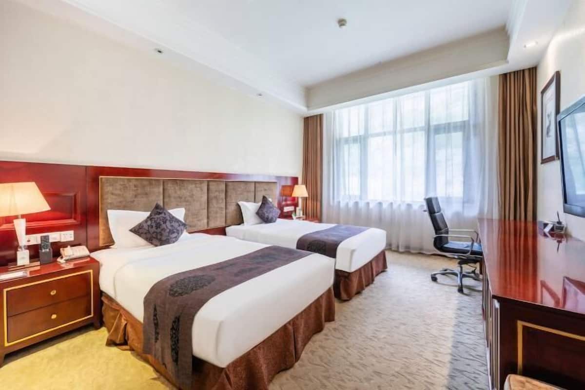 Quarto de hotel com duas camas, uma escrivaninha, uma cadeira e uma janela grande com cortinas.