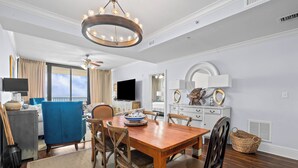 Comedor con mesa de madera, sillas, lámpara de araña, televisor y vista al balcón.