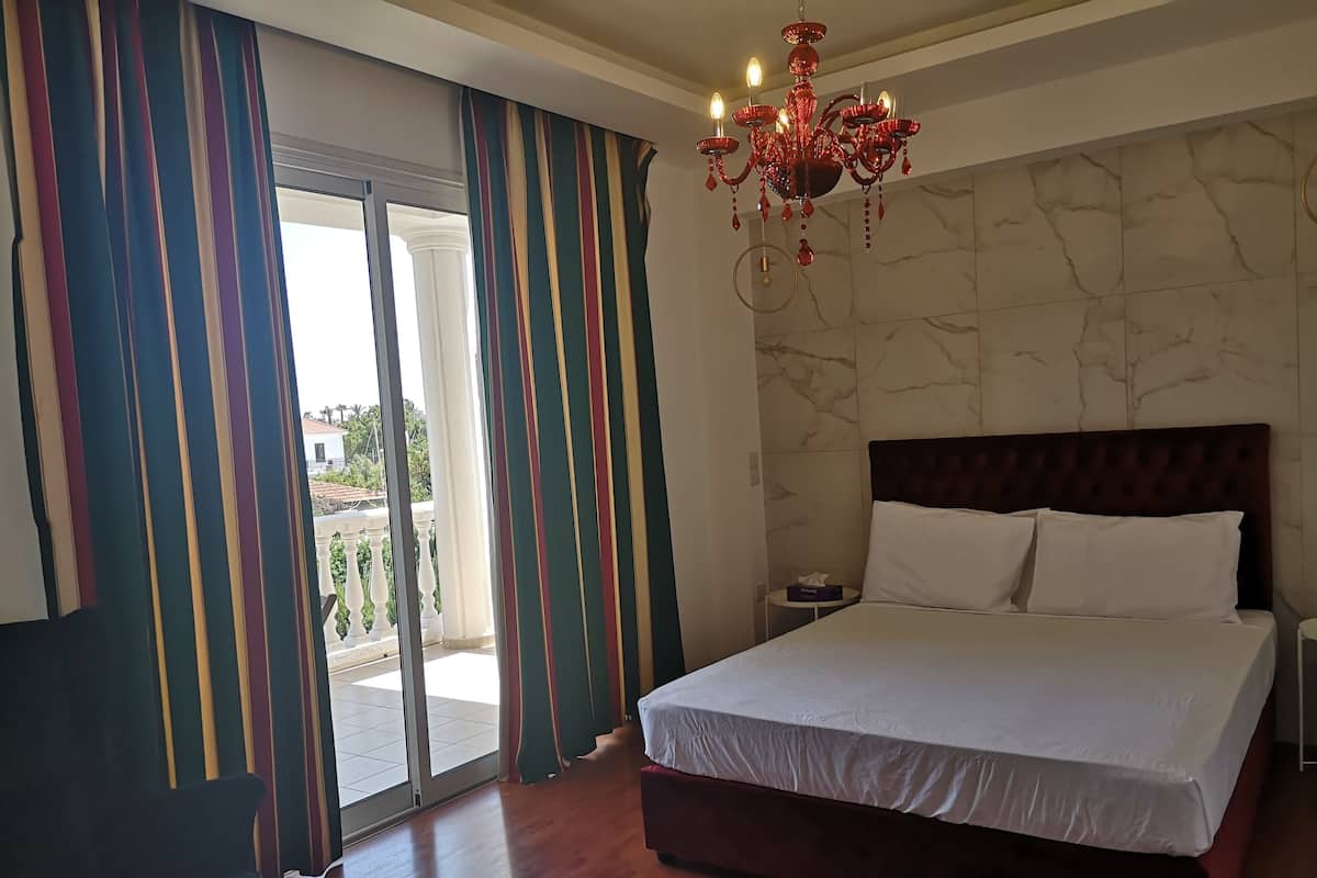 Habitación de hotel con una cama grande, una araña de cristal, vista a través de una puerta corrediza de vidrio y una pared de mármol.