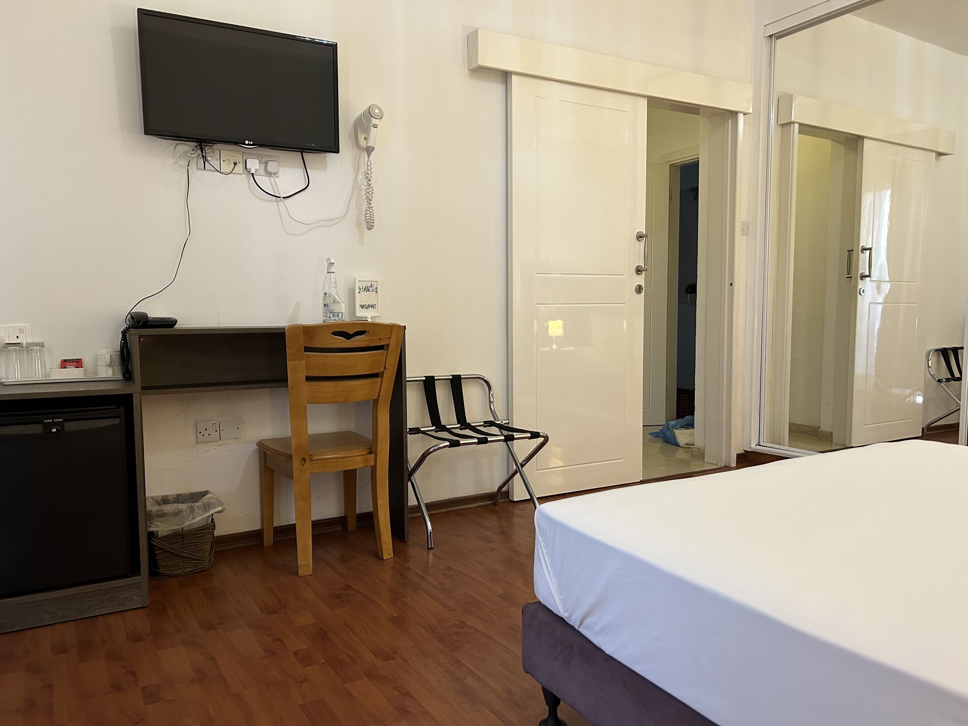 A hotel room with a bed, a desk, a chair, a TV mounted on the wall, and a door leading to another room.
