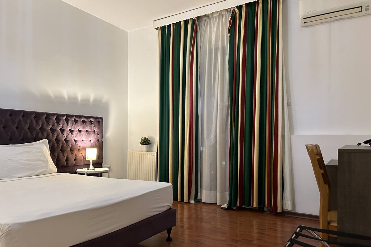 Habitación de hotel con una cama grande, una mesita de noche con lámpara, un radiador, una planta en maceta y un escritorio con silla.
