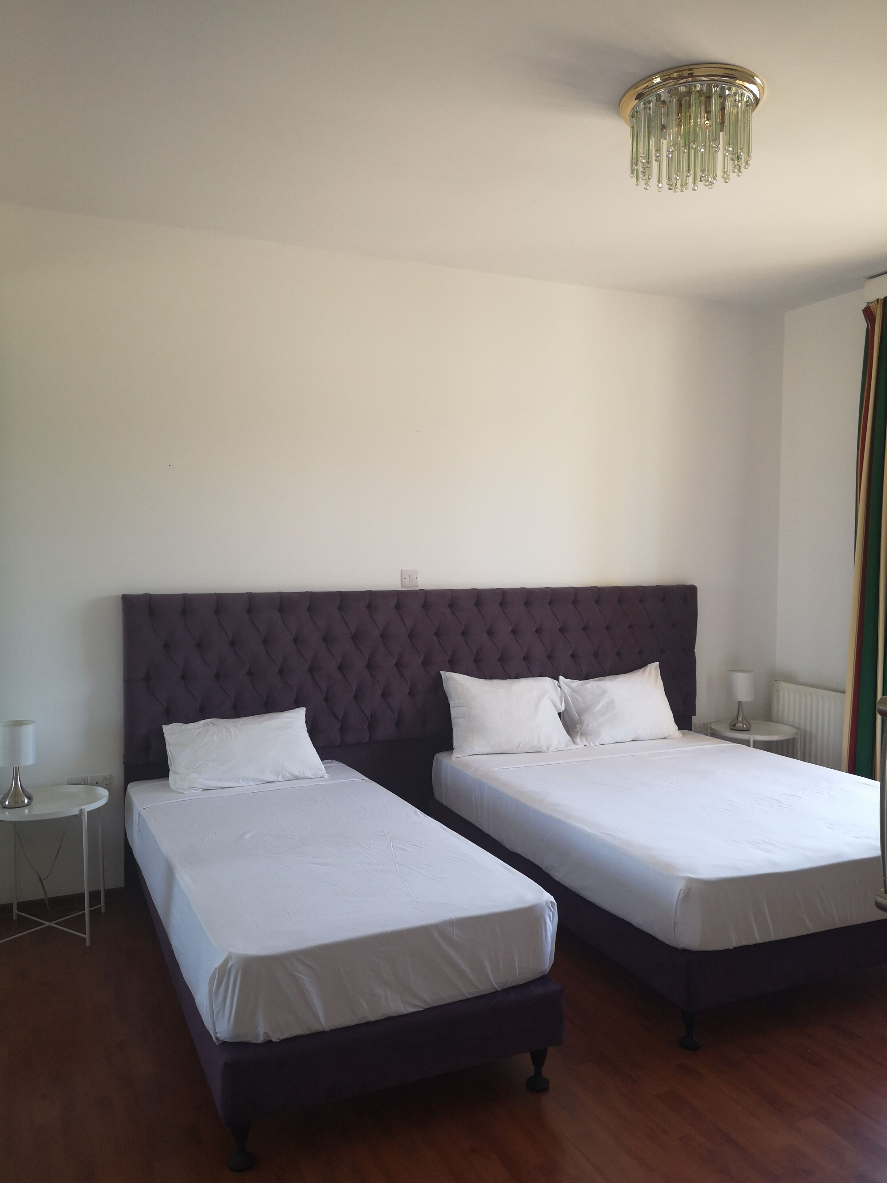 A room with two beds, a chandelier, a small table with a lamp, and a window with curtains.