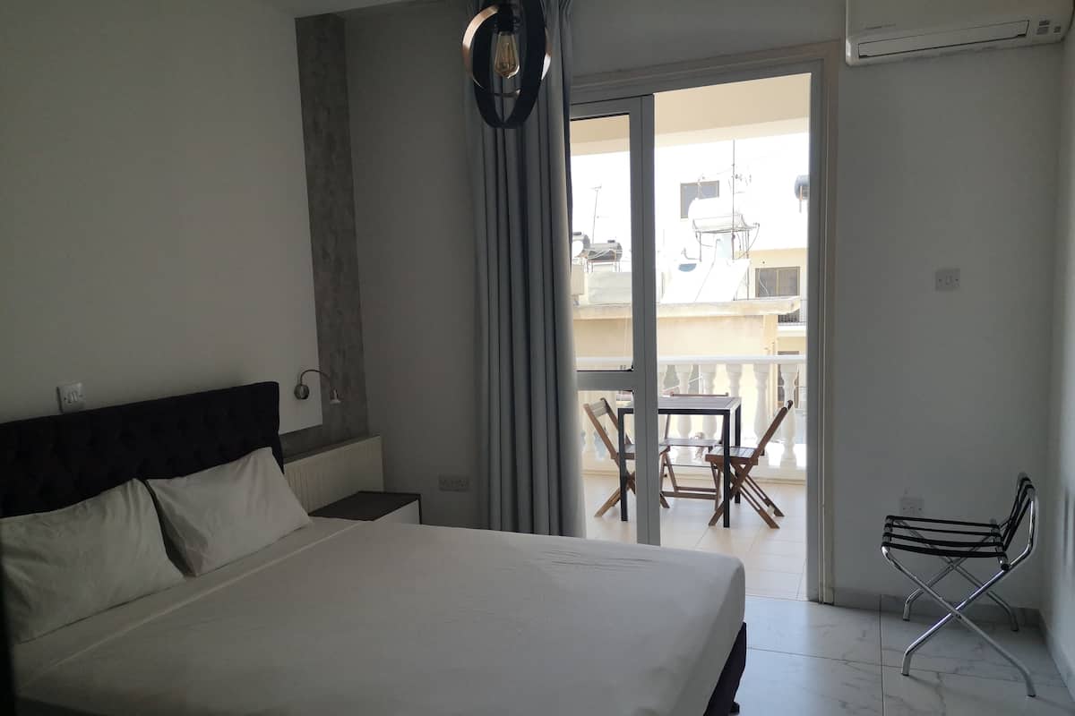 Habitación de hotel con una cama grande, una pequeña mesa con sillas y un balcón con vista al edificio.