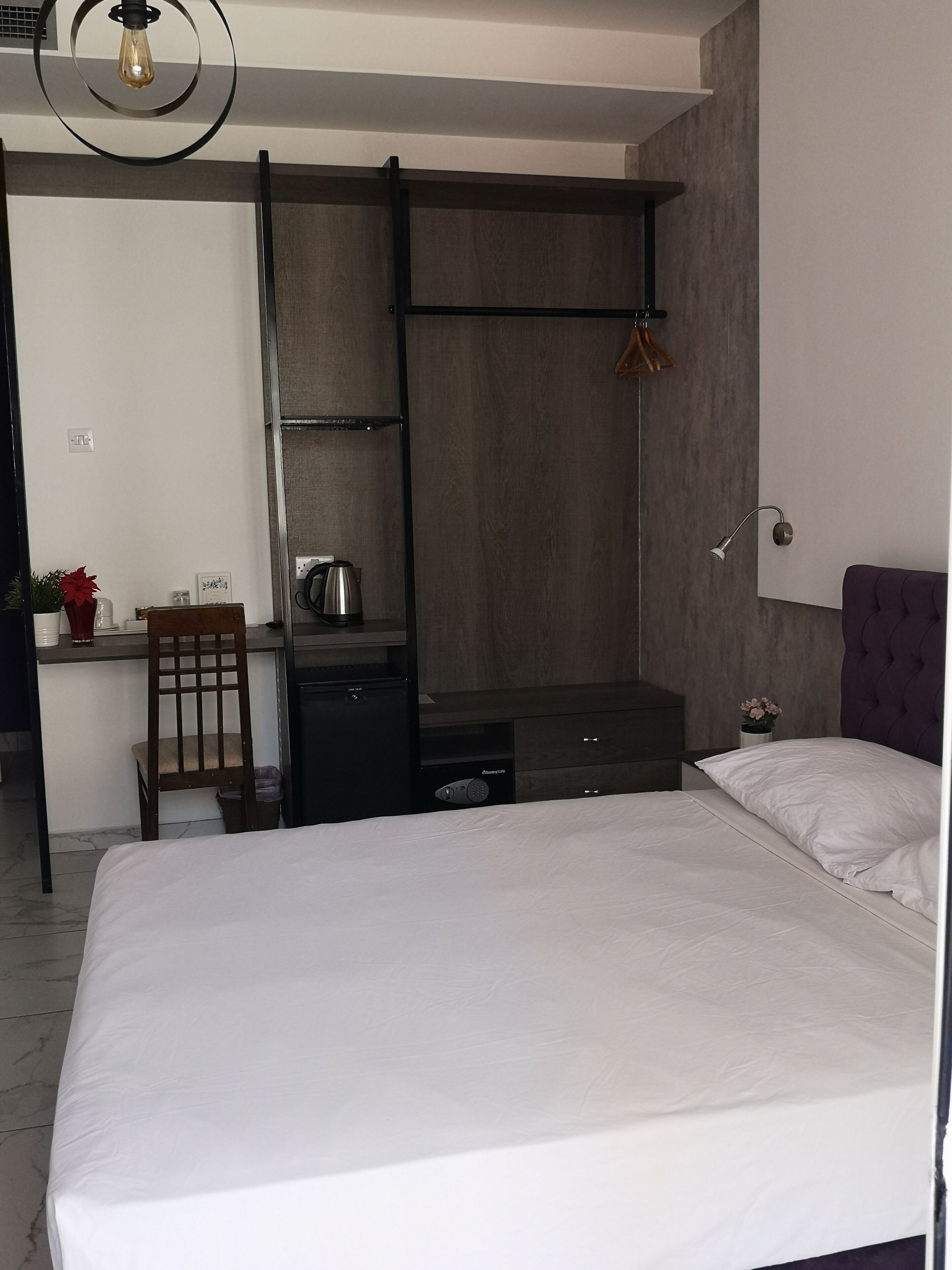 A bedroom with a bed, a desk, and a chair. There is a shelf unit with a kettle and a small plant.