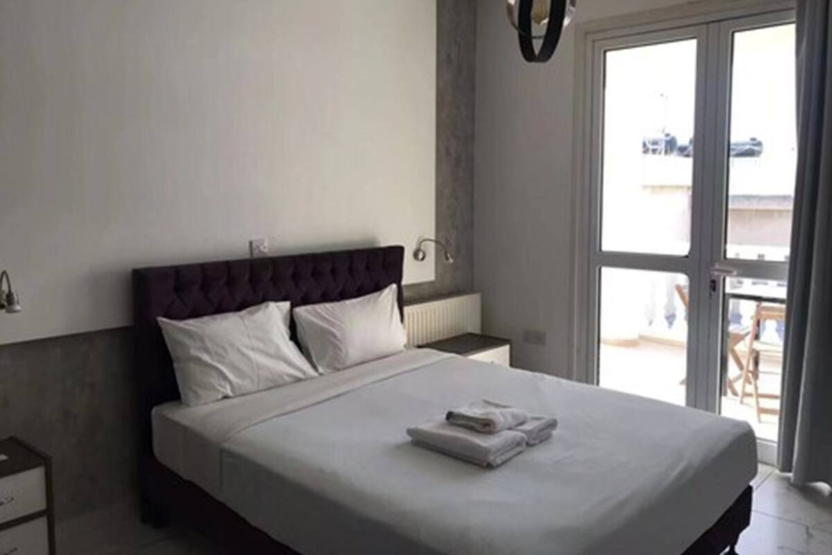 Un dormitorio con una cama grande, una mesita de noche, una lámpara de pared y vistas a través de un ventanal.