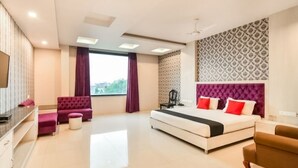 Una habitación de hotel moderna con una cama grande, una zona de estar con sofá y reposapiés, un televisor y un amplio ventanal con cortinas.