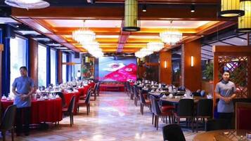 A restaurant with multiple tables and chairs, a large screen displaying a colorful image, and a ceiling with hanging lights.