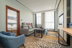 A modern hotel room with a sofa, a coffee table, a TV, and a large window with curtains.