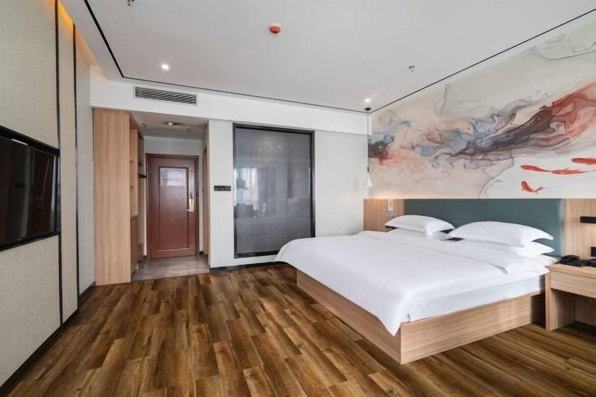 A modern hotel room with a wooden floor, a large bed with white linens, a desk with a chair, and a large abstract painting on the wall.