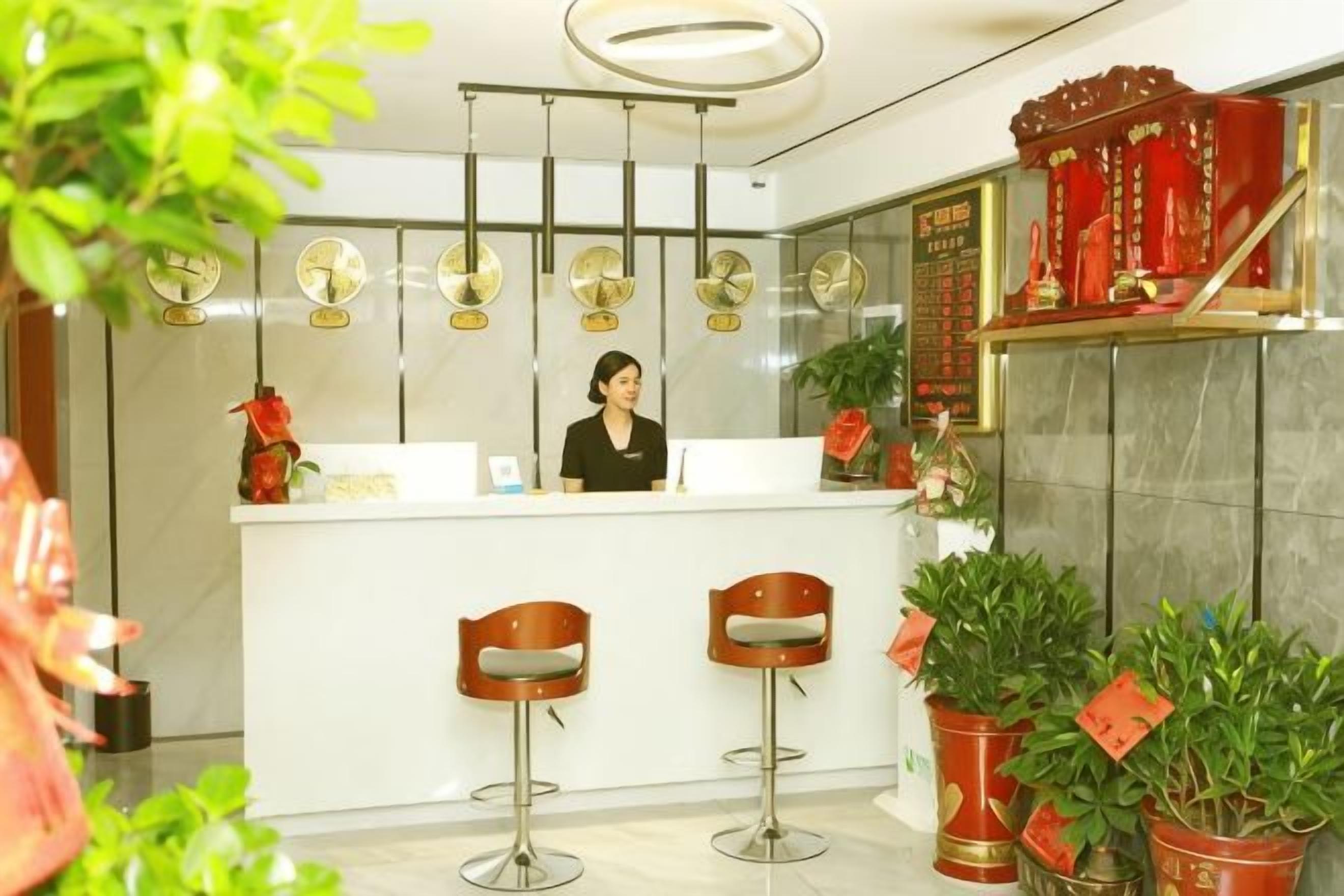 A reception area with a receptionist, a white counter, and three bar stools. There are potted plants and a decorative wall piece.