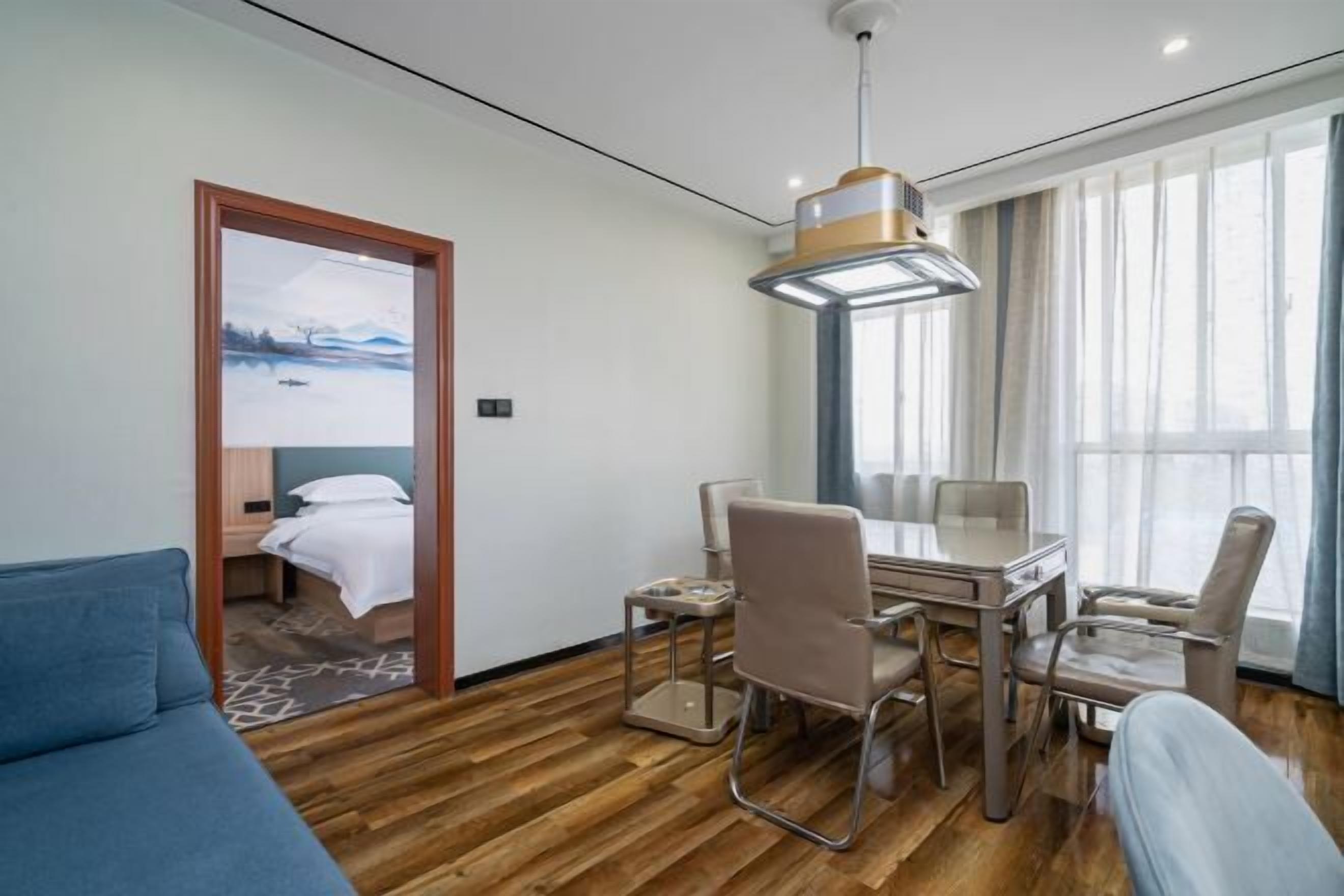 A hotel room with a wooden floor, a bed with white bedding, a desk with chairs, and a large window with curtains.