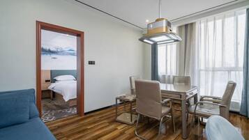A hotel room with a wooden floor, a bed with white bedding, a desk with chairs, and a large window with curtains.