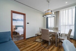 Habitación de hotel con piso de madera, cama con ropa blanca, escritorio y sillas, y un ventanal con cortinas.