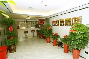 Zona de recepción de un hotel con mostrador, varias plantas en macetas y una pared con un cartel.