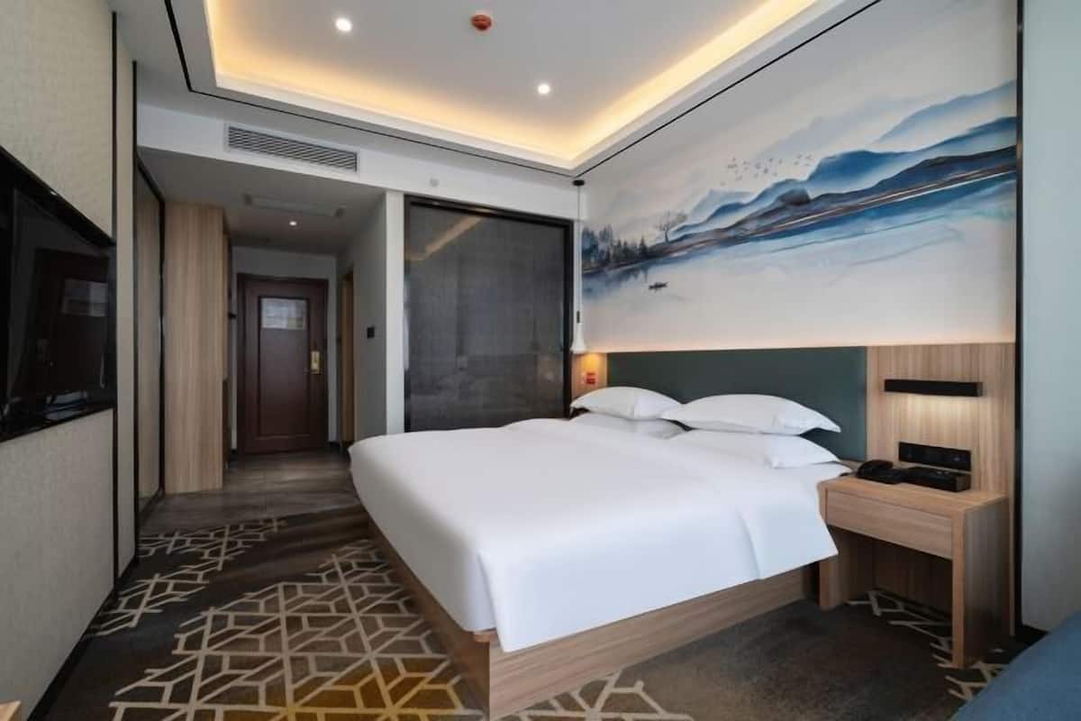 A modern hotel room with a large bed, a painting on the wall, and a desk with a lamp.