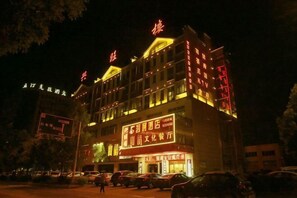 Um edifício com vários andares, sinalização iluminada, uma entrada destacada e área de estacionamento em frente.