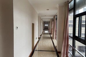 Un largo y estrecho pasillo de hotel con suelo de baldosas, ventanas amplias a un lado y una puerta al final.