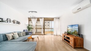 A modern living room with a sofa, a coffee table, a TV on a wooden stand, and a balcony with curtains. - MIKLAT Elegant Apt with Balcony & Sea View by FeelHome (Netanya)