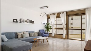 A living room with a grey sectional sofa, a small round table, and a blue armchair. There is a wall-mounted shelf with various items and a large window with curtains. - MIKLAT Elegant Apt with Balcony & Sea View by FeelHome (Netanya)
