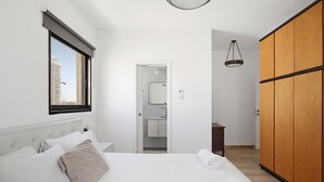 A hotel room with a bed, a wardrobe, a bathroom with a mirror, and a window with a view of buildings. - MIKLAT Elegant Apt with Balcony & Sea View by FeelHome (Netanya)
