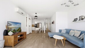 A modern living room with a sofa, a coffee table, and a TV. There is a dining area with a table and chairs. The room has wooden flooring and white walls. - MIKLAT Elegant Apt with Balcony & Sea View by FeelHome (Netanya)