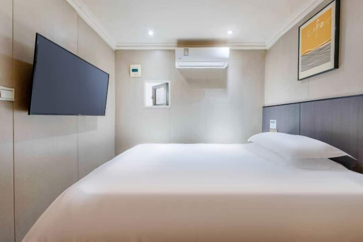 Ein Hotelzimmer mit Flachbildfernseher an der Wand, einem Bett mit weißer Bettwäsche, einer Klimaanlage und einem gerahmten Bild.