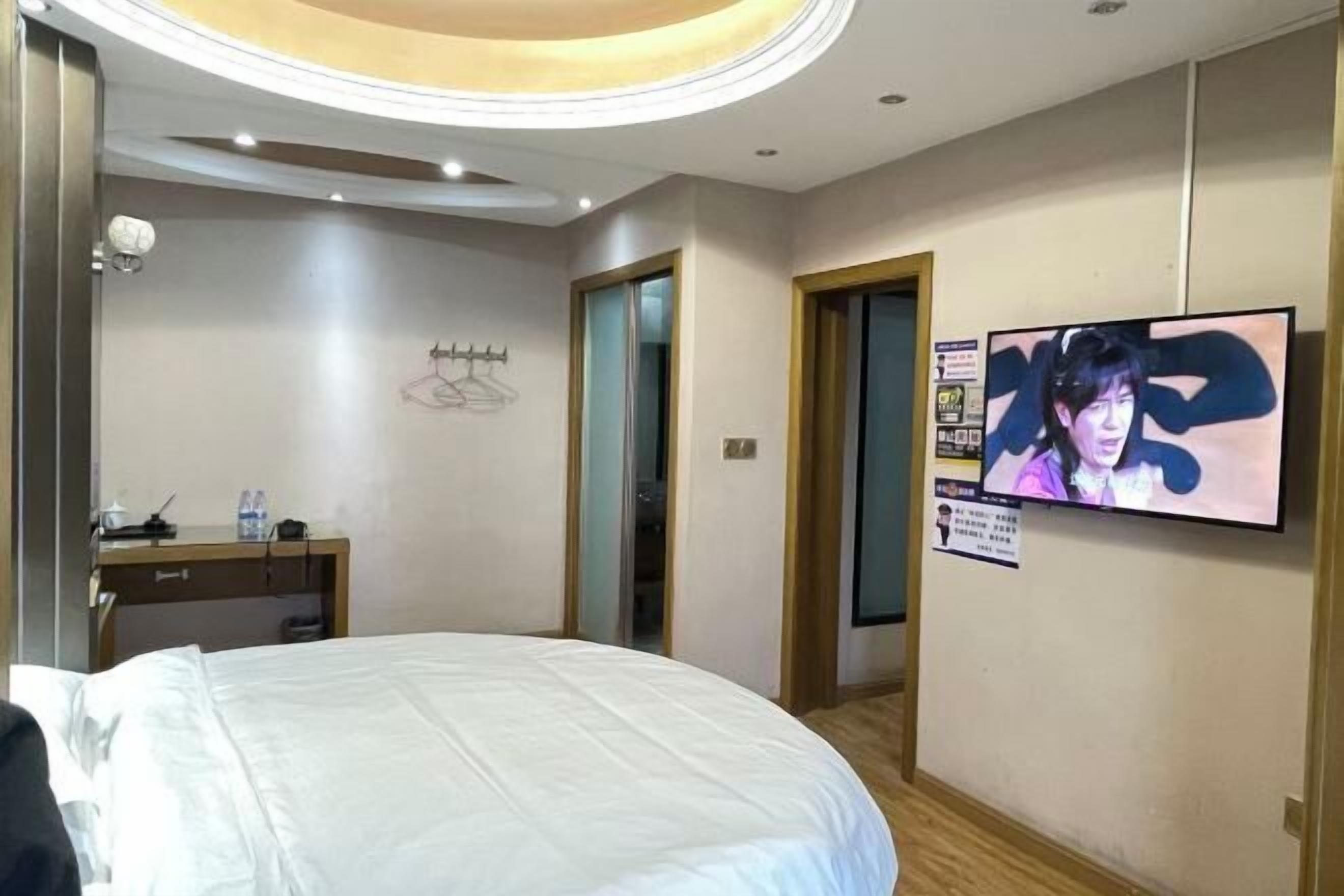 Ein Hotelzimmer mit einem Bett, einem Schreibtisch mit Stuhl, einem an der Wand befestigten Fernseher und einer Tür zu einem weiteren Raum.