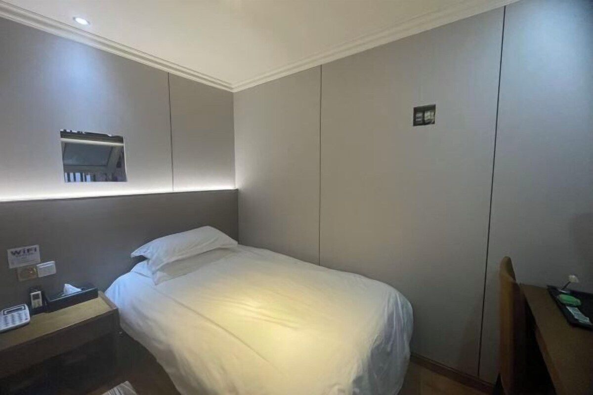 Ein Hotelzimmer mit einem Einzelbett, einem Nachttisch mit Lampe, einem kleinen Fenster und einer wandmontierten Steuerung.