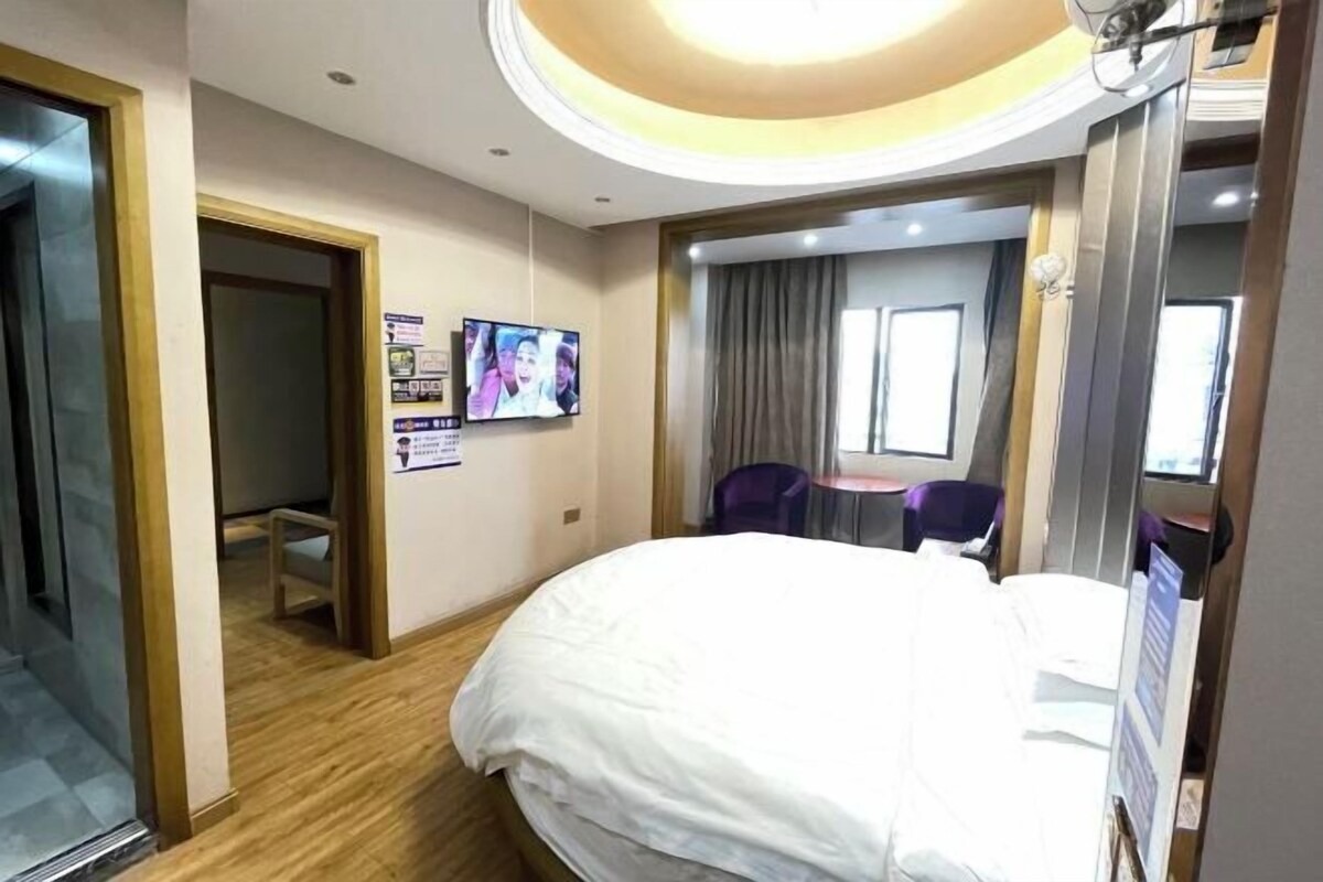 Ein Hotelzimmer mit einem großen Bett, einem an der Wand befestigten Fernseher, einem kleinen Tisch mit Stühlen und einem Fenster mit Vorhängen.