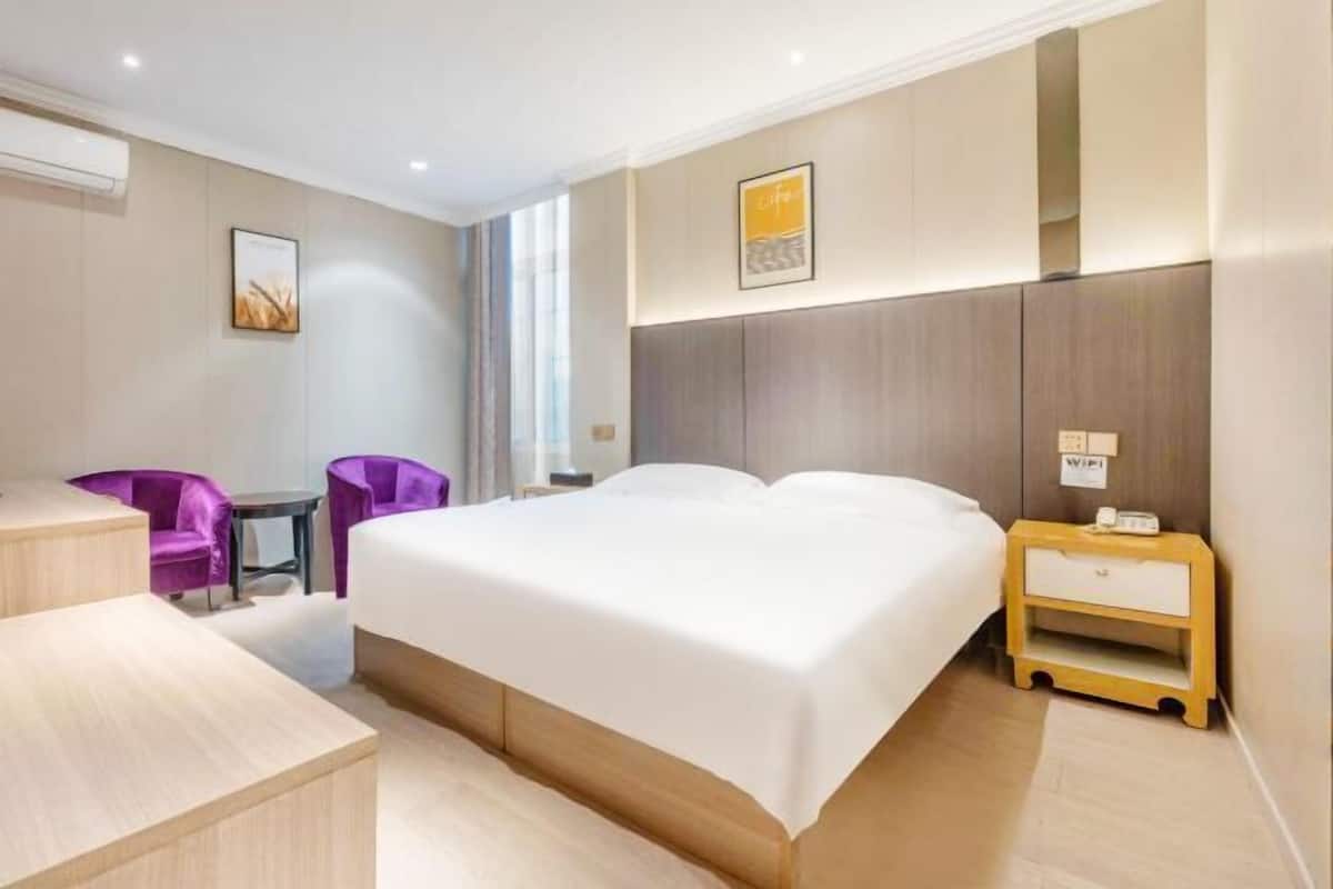 Ein Hotelzimmer mit einem Bett, einem Schreibtisch, zwei violetten Stühlen, einem kleinen Tisch, einem Nachttisch mit Telefon und einem Bild an der Wand.