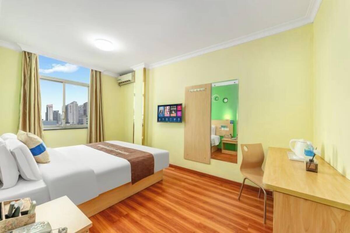 A hotel room with a bed, a desk, a chair, and a view of the cityscape through the window.