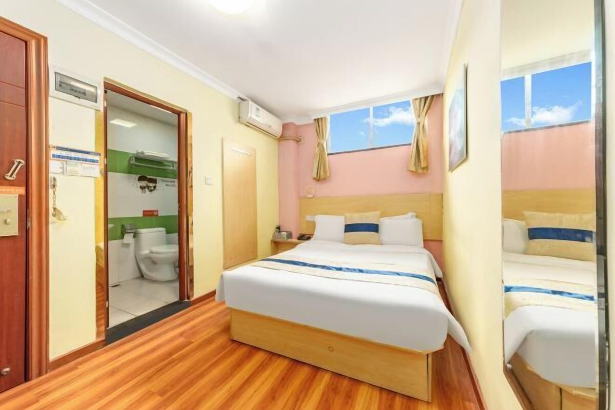 A hotel room with a wooden floor, a bed with white bedding and blue accents, a window with curtains, and a door leading to a bathroom.
